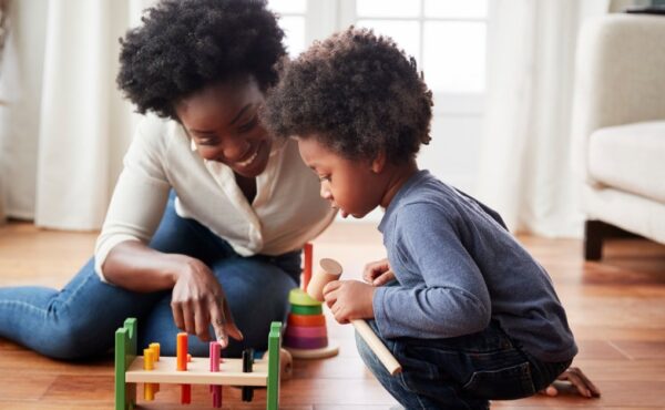 A mom and son playing with a wooden block toy on the floor of their home. The little boy is crouched and looking at the toy.