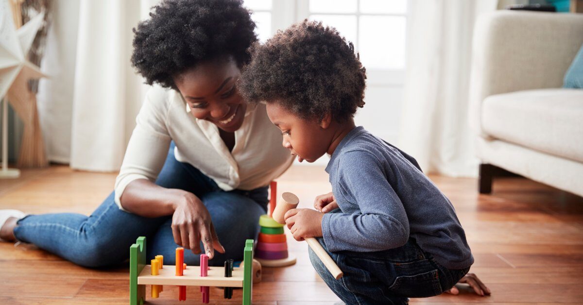 A mom and son playing with a wooden block toy on the floor of their home. The little boy is crouched and looking at the toy.