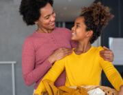 A mother and daughter are smiling at one another as the daughter holds an organized basket with laundry.