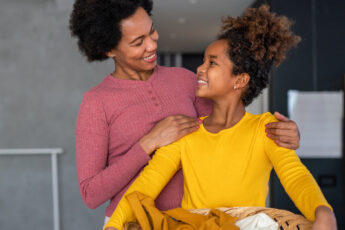 A mother and daughter are smiling at one another as the daughter holds an organized basket with laundry.