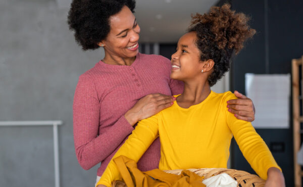 A mother and daughter are smiling at one another as the daughter holds an organized basket with laundry.