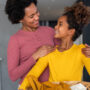 A mother and daughter are smiling at one another as the daughter holds an organized basket with laundry.