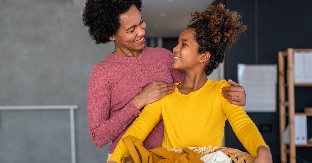 A mother and daughter are smiling at one another as the daughter holds an organized basket with laundry.