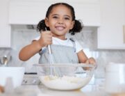 A happy toddler at the counter with baking supplies. She is using a whisk to stir the ingredients in the mixing bowl.