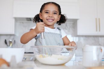 A happy toddler at the counter with baking supplies. She is using a whisk to stir the ingredients in the mixing bowl.