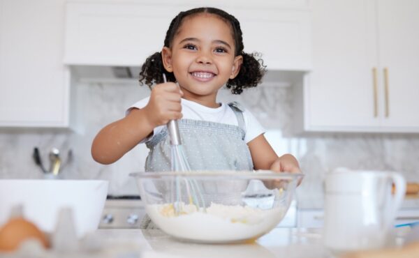 A happy toddler at the counter with baking supplies. She is using a whisk to stir the ingredients in the mixing bowl.