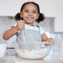 A happy toddler at the counter with baking supplies. She is using a whisk to stir the ingredients in the mixing bowl.