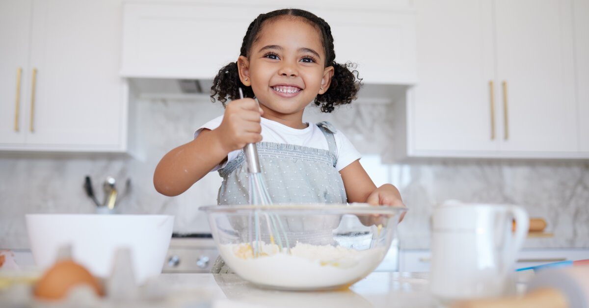 A happy toddler at the counter with baking supplies. She is using a whisk to stir the ingredients in the mixing bowl.