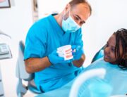 A pediatric dentist showing a set of fake teeth to a young Black girl. She is sitting in the chair with an apron on.