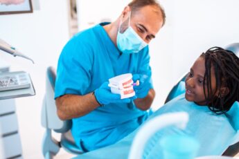 A pediatric dentist showing a set of fake teeth to a young Black girl. She is sitting in the chair with an apron on.