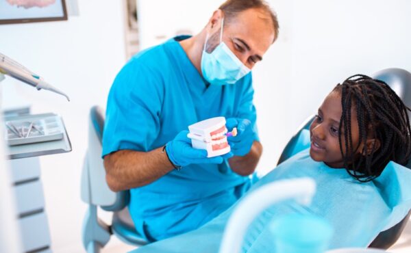 A pediatric dentist showing a set of fake teeth to a young Black girl. She is sitting in the chair with an apron on.