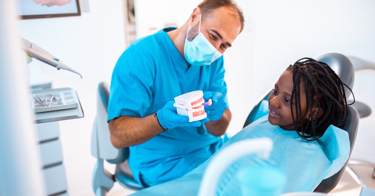 A pediatric dentist showing a set of fake teeth to a young Black girl. She is sitting in the chair with an apron on.