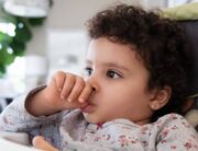 A cute toddler sitting in her high chair as she sucks on their thumb. She's wearing a gray printed top and has curly hair.