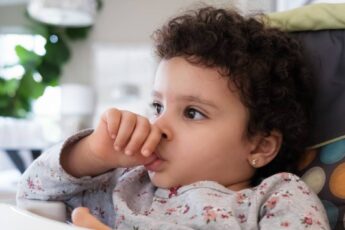 A cute toddler sitting in her high chair as she sucks on their thumb. She's wearing a gray printed top and has curly hair.