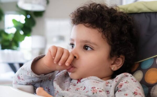 A cute toddler sitting in her high chair as she sucks on their thumb. She's wearing a gray printed top and has curly hair.