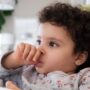 A cute toddler sitting in her high chair as she sucks on their thumb. She's wearing a gray printed top and has curly hair.