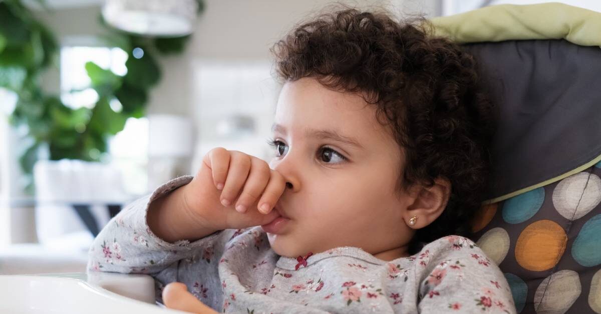 A cute toddler sitting in her high chair as she sucks on their thumb. She's wearing a gray printed top and has curly hair.