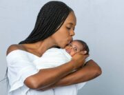 A Black mother with braids kisses her newborn on the forehead as she cradles him. She wears a white dress.