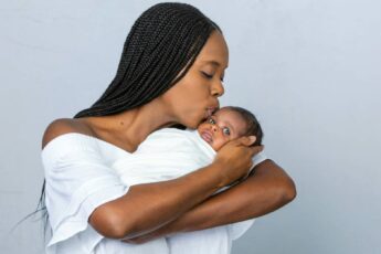 A Black mother with braids kisses her newborn on the forehead as she cradles him. She wears a white dress.