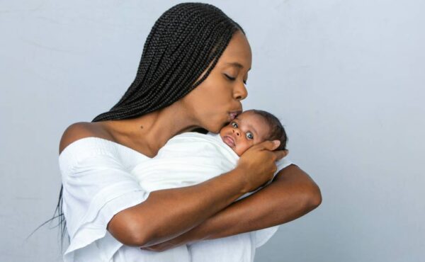 A Black mother with braids kisses her newborn on the forehead as she cradles him. She wears a white dress.