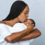 A Black mother with braids kisses her newborn on the forehead as she cradles him. She wears a white dress.