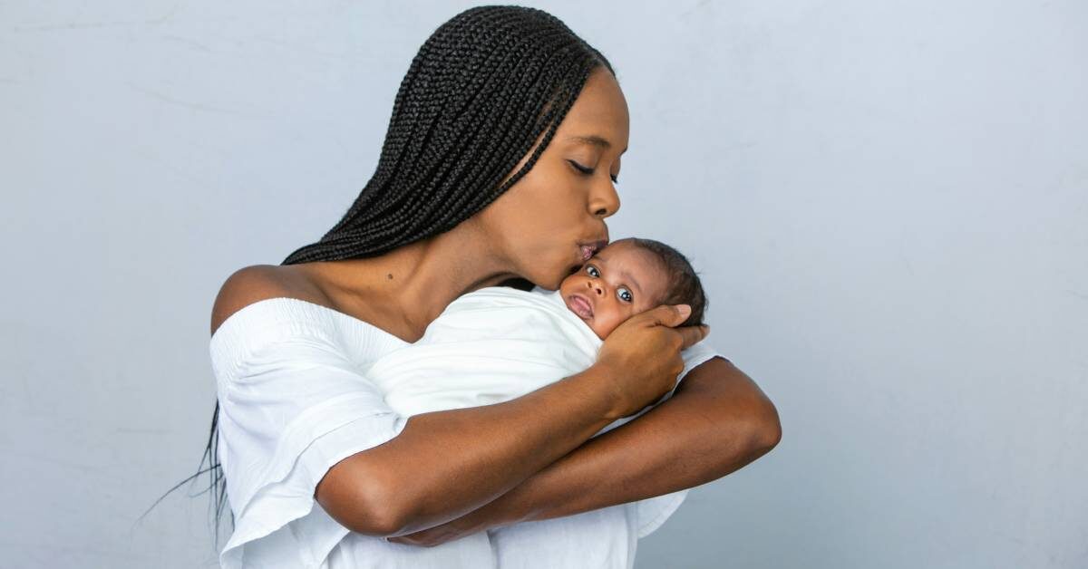 A Black mother with braids kisses her newborn on the forehead as she cradles him. She wears a white dress.