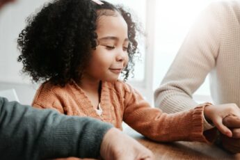 A little girl with curly hair is seated at a table and holding hands with two adults. Her eyes are closed, and she smiles.