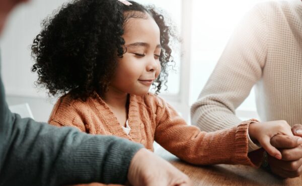 A little girl with curly hair is seated at a table and holding hands with two adults. Her eyes are closed, and she smiles.