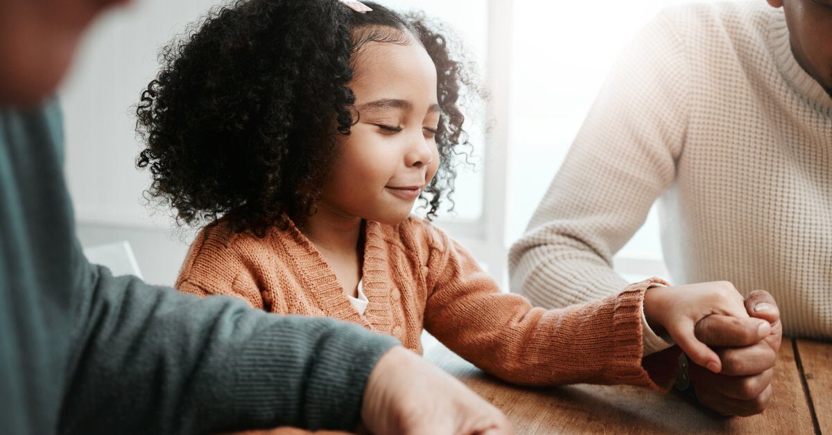 A little girl with curly hair is seated at a table and holding hands with two adults. Her eyes are closed, and she smiles.