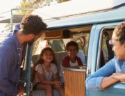 A Black family of four on an RV road trip. The mom is in the passenger seat, the kids in the back, and the dad by the door.