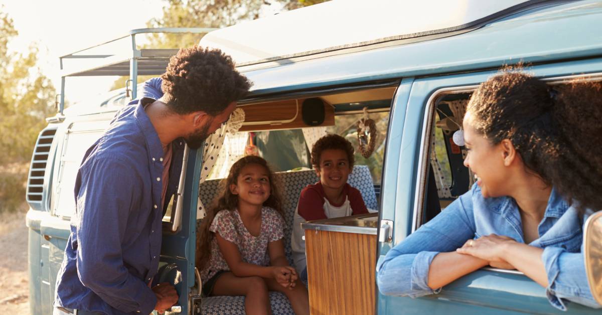 A Black family of four on an RV road trip. The mom is in the passenger seat, the kids in the back, and the dad by the door.