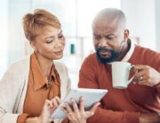 A black man and woman sit at a table and look at an electronic tablet while drinking coffee and inspecting bills.