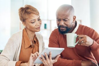 A black man and woman sit at a table and look at an electronic tablet while drinking coffee and inspecting bills.