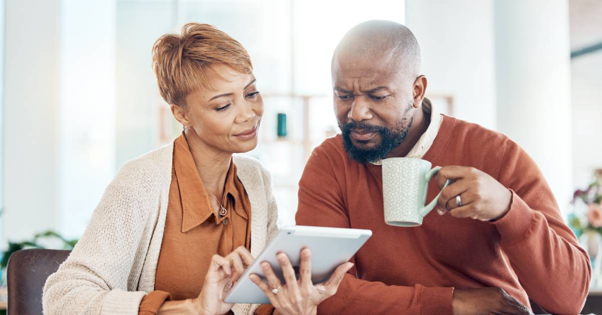 A black man and woman sit at a table and look at an electronic tablet while drinking coffee and inspecting bills.