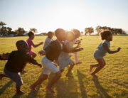 A group of Black children play and run together across an open field. The sun shines in the background.