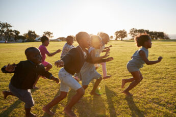 A group of Black children play and run together across an open field. The sun shines in the background.