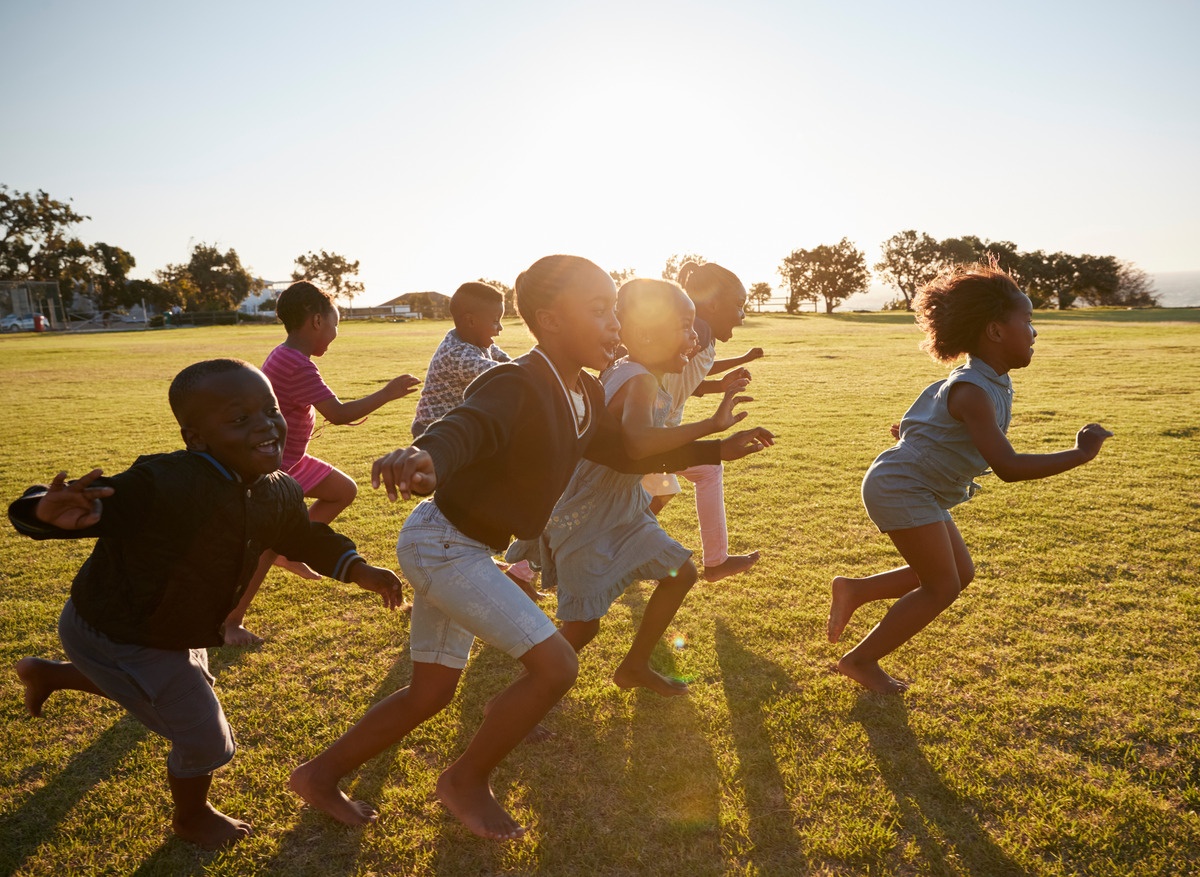 A group of Black children play and run together across an open field. The sun shines in the background.