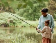 A Black mother stands beside her young child and guides them as they hold a long fishing rod out toward the water.