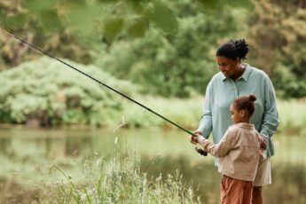 A Black mother stands beside her young child and guides them as they hold a long fishing rod out toward the water.