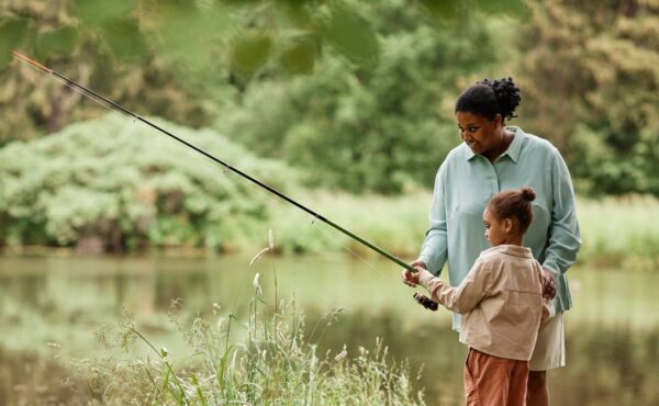 A Black mother stands beside her young child and guides them as they hold a long fishing rod out toward the water.