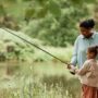 A Black mother stands beside her young child and guides them as they hold a long fishing rod out toward the water.