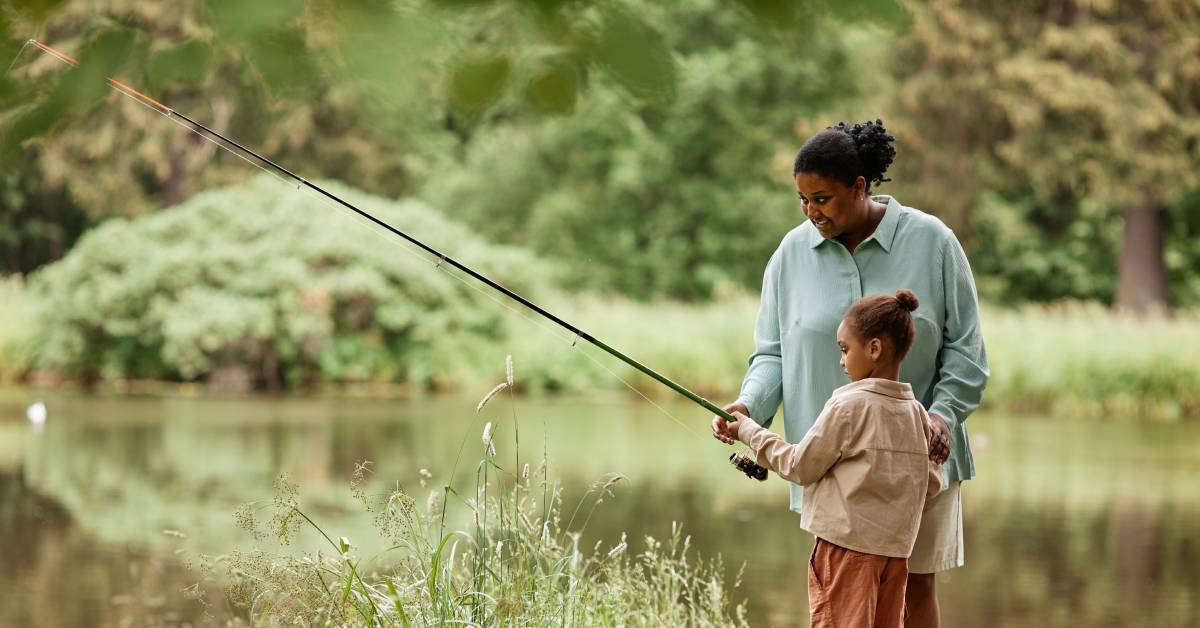 A Black mother stands beside her young child and guides them as they hold a long fishing rod out toward the water.