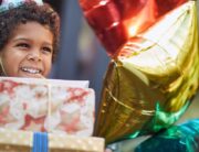 A young Black child with a stack of birthday gifts in his hands. He's smiling and wearing a birthday party hat.