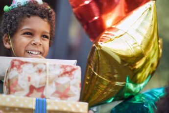 A young Black child with a stack of birthday gifts in his hands. He's smiling and wearing a birthday party hat.