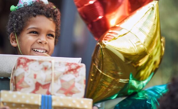 A young Black child with a stack of birthday gifts in his hands. He's smiling and wearing a birthday party hat.