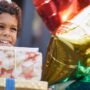 A young Black child with a stack of birthday gifts in his hands. He's smiling and wearing a birthday party hat.