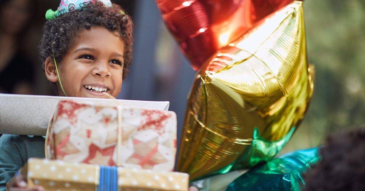 A young Black child with a stack of birthday gifts in his hands. He's smiling and wearing a birthday party hat.