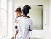 A Black mother holding her child near the sink of a clean white bathroom. They are both wearing pajama outfits.