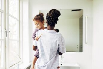 A Black mother holding her child near the sink of a clean white bathroom. They are both wearing pajama outfits.