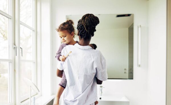 A Black mother holding her child near the sink of a clean white bathroom. They are both wearing pajama outfits.